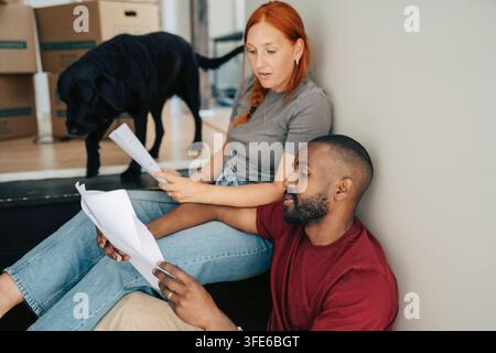 Verschiedenes Paar betrachtet Papierkram mit Haustierhund im Hintergrund während des Umzugs Stockfoto