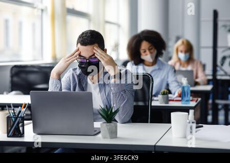 Millennial-Mann mit Schutzmaske stützt seinen Kopf auf die Hände, sitzt am Tisch und schaut auf den Laptop Stockfoto