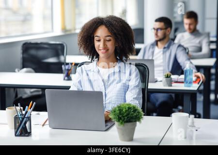 Mitarbeiter arbeitet während des Coronavirus im Coworking Office. Lächelnde afroamerikanische tausendjährige Frau, tippen auf Laptop am Arbeitsplatz mit antiseptischen Stockfoto