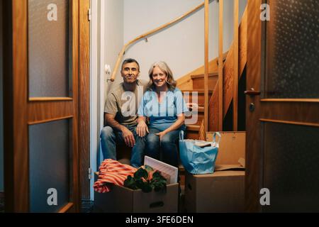 Porträt eines lächelnden Paares, das auf Holztreppen sitzt, umgeben von Umzugskartons Stockfoto