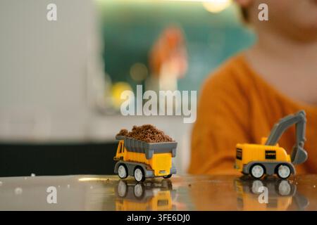 Gelbe Spielzeugkipplaster mit Dreck auf glänzender Tischoberfläche im Nahaufnahme. Bauspielzeuge gefüllt mit brauner Erde, die drinnen fantasievolles Spielen ermöglichen. Bunte Spielfahrzeuge für Kinder, die Spaß im Sandkasten zu Hause haben Stockfoto