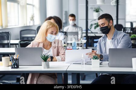 Millennial-Mann und -Frau mit Papieren am Arbeitsplatz mit Laptops und einer Flasche Desinfektionsmittel sprechen nach der Quarantäne Stockfoto