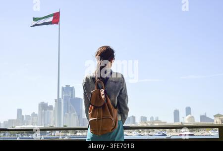 Ein Tourist mit einem Rucksack bewundert die Stadtlandschaft von Flag Island in Sharjah. Rückansicht einer Frau auf dem Damm und die Flagge der VAE Stockfoto