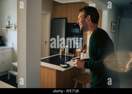 Seitenansicht eines glücklichen Mannes, der eine Kaffeetasse in seiner Küche zu Hause hält Stockfoto