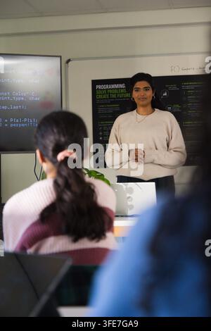 Indische Moderatorin in beigem Pullover präsentiert eine Diskussion, während Studenten Code auf dem Bildschirm betrachten Stockfoto