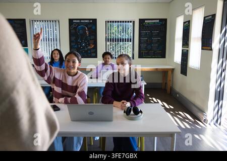 Schülerinnen heben die Hand und nutzen Laptops in einem Computerraum mit Postern und Kopfhörern Stockfoto