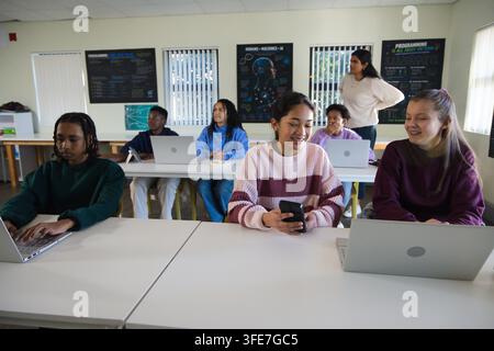 Verschiedene Klassenkameraden verwenden Laptops und Handys im Computerraum mit einer Lehrerin, die in der Nähe von Postern beaufsichtigt Stockfoto