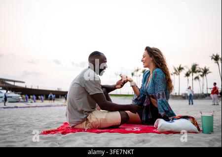 Zwangloses junges Paar, das ein spontanes abendliches Picknick auf dem Sand genießt, das sich auf ihren entspannten Stil konzentriert Stockfoto