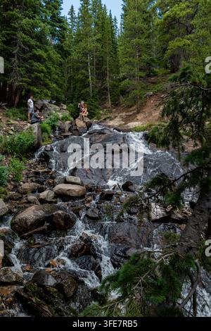 Rocky-Mountain-Wald - Juli 2022: Ein Wasserfall stürzt über moosbedeckte Felsbrocken in einem Kiefernwald, Wanderer ruhen auf Felsen nahe dem Gipfel des Wasserfalls Stockfoto
