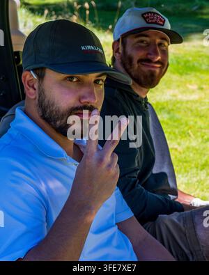 Zwei lächelnde Männer fahren gemeinsam in einem Golfwagen auf einem sonnigen Berggolfplatz, einer zeigt ein Peace-Zeichen. Stockfoto