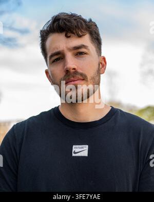 Ein junger bärtiger Mann schaut direkt in die Kamera in einem Outdoor-Porträt, trägt ein dunkles sportliches T-Shirt. Stockfoto