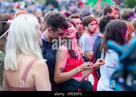 JUNGES PAAR UMGARMT SICH AUF MUSIKFESTIVAL, REPORTAGE, GREEN MAN FESTIVAL 2018, BRECON, WALES, UK, 2018: Ein junger Mann umarmt eine lächelnde Frau von hinten, während sie sich inmitten einer großen Menge auf einem Open-Air-Musikfestival amüsieren, beleuchtet von rosa und blauen Lichtern. Foto: Rob Watkins. INFO: Das Green Man Festival begann 2003 in Wales. Es findet jährlich an dem dritten Wochenende im August in den Brecon Beacons statt und feiert alternative Musik, Kunst, Literatur, Wissenschaft und familienfreundliche Kultur in einer wunderschönen Berglandschaft. Stockfoto