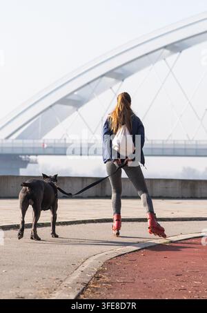 Frau rollert mit einem großen schwarzen Hund an der Leine entlang einer Uferpromenade an einem sonnigen Tag und kombiniert dabei Freizeit, Sport und Gesellschaft Stockfoto