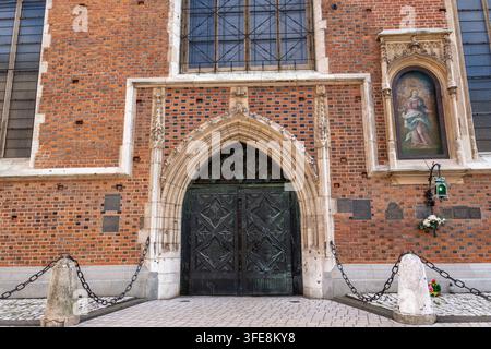 Krakau, Polen – 29.08.2026 Der kunstvolle Kircheneingang steht in einer roten Backsteinmauer mit spitzem Steinbogen und dunkler Metalltür. Gedenktafeln, buntes Glas Stockfoto