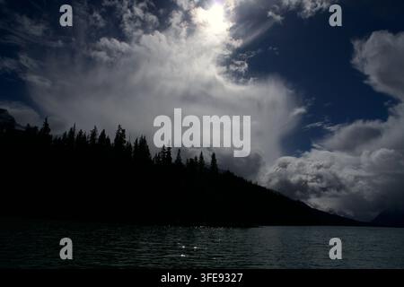 Helle Sonne scheint durch dramatische Gewitterwolken über einer dunklen Silhouette eines Kiefernwaldes an einem breiten Alpelsee Stockfoto
