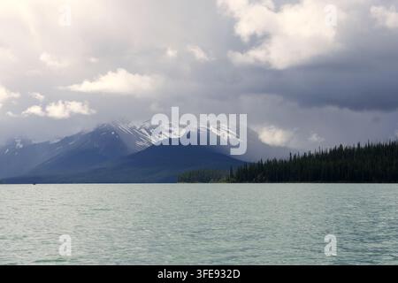 Ruhiges Algensee unter einem dramatischen stürmischen Himmel mit dunklen Wolken, die über entfernte schneebedeckte Berggipfel ziehen Stockfoto