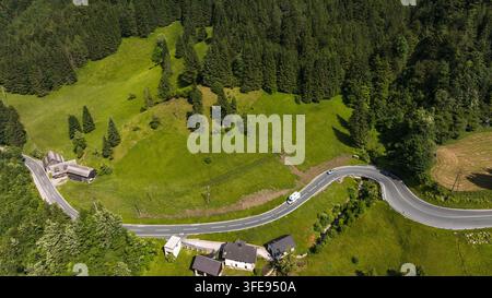 Drohne fängt die gewundene Asphaltstraße durch das üppige grüne Bergtal in Österreich ein. Traditionelle Häuser liegen am Waldrand während des sonnigen Sommertages in der Alpenregion nahe der Grenze. Stockfoto