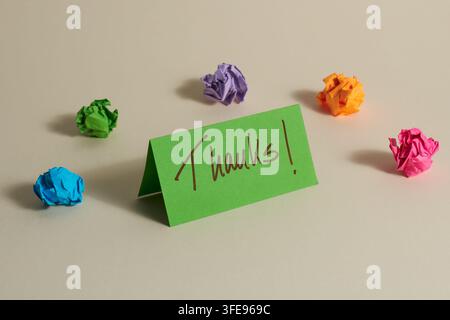 Eine grüne gefaltete Karte mit einer handgeschriebenen Nachricht "Danke!" umgeben von bunten zerknüllten Papierbällen auf einem neutralen Hintergrund. Die fröhliche Komposition Stockfoto