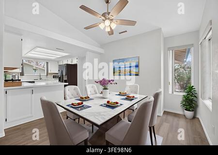 Luftiger weißer Innenraum mit Marmoresstisch, getufteten Stühlen, Deckenventilator mit Lichtern und großen Fenstern, die warmes Sonnenlicht für die Mahlzeiten des Alltags hereinlassen Stockfoto