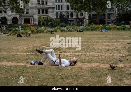 Ein Stadtarbeiter liegt auf dem Gras im Finsbury Square EC2 und hört Musik über Kopfhörer, die an sein Smartphone gebunden sind, City of London, England, UK. Stockfoto