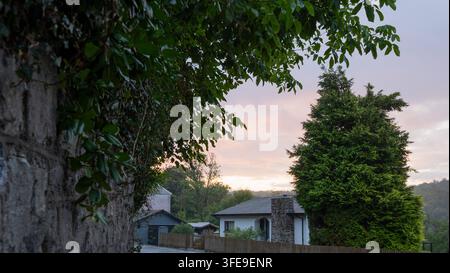 Vorstadthaus-Außenansicht idyllischer Blick auf ein Wohngebäude mit Steinkamin, Holzzaun, immergrünem Baum und efeubedeckter Steinmauer im Vordergrund Stockfoto