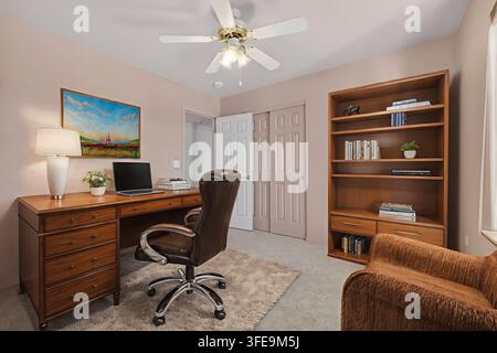 Gemütliche Innenraum-Home-Office-Szene mit Holzschreibtisch und Laptop, Bücherstapel, hohem Bücherregal, Lampe und Pflanze, Deckenventilator und gerahmter Landschaftswandkunst Stockfoto