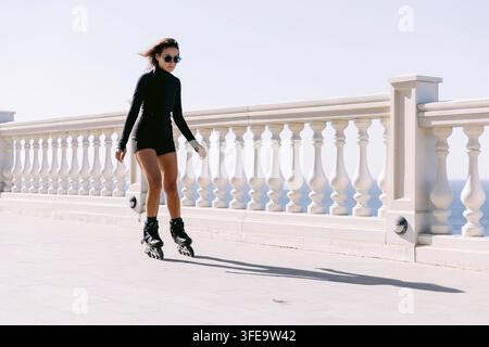 Frau in schwarzer Sportkleidung rollert auf einer sonnigen Uferpromenade Stockfoto