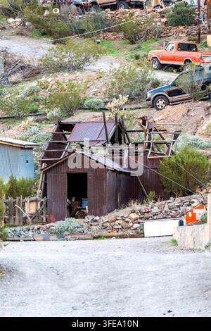 Ein verwitterter Holzschuppen befindet sich auf einem felsigen Hang mit einem gewellten Metalldach, umgeben von Baumstämmen, Steinen und alten Fahrzeugen einschließlich eines kleinen orangen trucks Stockfoto