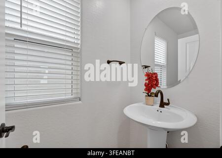 Saubere moderne Badezimmer-Vignette mit weißem Standwaschbecken, Chrom-Wasserhahn, großem kreisförmigem Spiegel, Fensterrollos und roter Orchidee im Vase, die Ruhe bringt Stockfoto