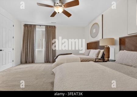 Helles Zimmer im Hotelstil mit zwei Betten mit beigefarbener Bettwäsche, einem hölzernen Kopfteil und passenden Nachttischen. Ein Deckenventilator mit einem hellen, weichen Teppich Stockfoto