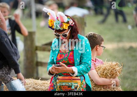 FRAU IN EINEM KLEINLICHEN HASENOHREN-OUTFIT AUF EINEM FESTIVAL, REPORTAGE, GREEN MAN FESTIVAL 2019, BRECON, WALES, UK, 2019: Eine lächelnde Frau, die eine bunte Jacke, eine herzförmige Sonnenbrille und ein Blumenstirnband mit Hasenohren trägt, schaut auf ihr Handy. Foto: Rob Watkins. INFO: Das Green Man Festival begann 2003 in Wales. Es findet jährlich an dem dritten Wochenende im August in den Brecon Beacons statt und feiert alternative Musik, Kunst, Literatur, Wissenschaft und familienfreundliche Kultur in einer wunderschönen Berglandschaft. Stockfoto