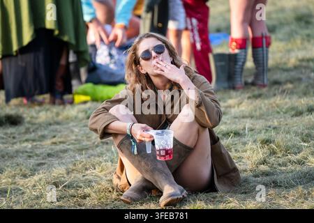 FESTIVALBESUCHER RAUCHT UND ENTSPANT SICH AUF DER WIESE, REPORTAGE, GREEN MAN FESTIVAL 2019, BRECON, WALES, UK, 2019: Eine junge Frau mit Sonnenbrille und brauner Jacke sitzt auf der Wiese, schaut zur Seite, während sie eine Zigarette raucht und einen Plastikbecher hält. Foto: Rob Watkins. INFO: Das Green Man Festival begann 2003 in Wales. Es findet jährlich im dritten Augustwochenende in den Brecon Beacons statt und feiert alternative Musik, Kunst, Literatur, Wissenschaft und familienfreundliche Kultur in einer wunderschönen Berglandschaft. Stockfoto