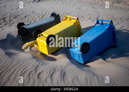 Drei bunte Kunststoff-Recyclingbehälter liegen auf den Seiten in den sandigen Stranddünen. Umgestürzte Müllcontainer, die Sturmschäden oder Abfallmanagement betonen Stockfoto