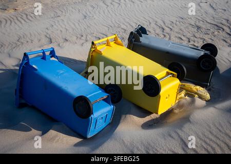 Drei bunte Kunststoff-Recyclingbehälter liegen auf den Seiten in den sandigen Stranddünen. Umgestürzte Müllcontainer, die Sturmschäden oder Abfallmanagement betonen Stockfoto