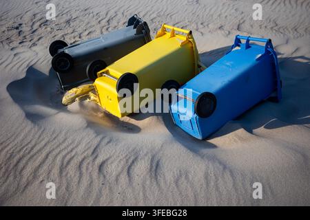 Drei bunte Kunststoff-Recyclingbehälter liegen auf den Seiten in den sandigen Stranddünen. Umgestürzte Müllcontainer, die Sturmschäden oder Abfallmanagement betonen Stockfoto