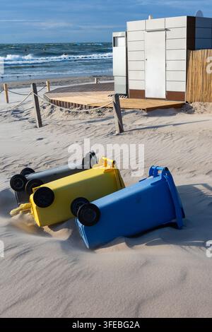 Drei bunte Kunststoff-Recyclingbehälter liegen auf den Seiten in den sandigen Stranddünen. Umgestürzte Müllcontainer, die Sturmschäden oder Abfallmanagement betonen Stockfoto