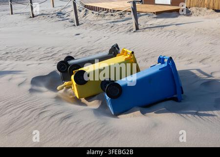 Drei bunte Kunststoff-Recyclingbehälter liegen auf den Seiten in den sandigen Stranddünen. Umgestürzte Müllcontainer, die Sturmschäden oder Abfallmanagement betonen Stockfoto
