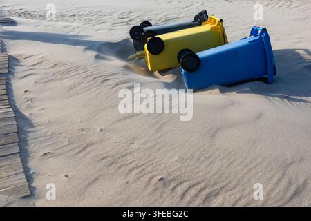 Drei bunte Kunststoff-Recyclingbehälter liegen auf den Seiten in den sandigen Stranddünen. Umgestürzte Müllcontainer, die Sturmschäden oder Abfallmanagement betonen Stockfoto