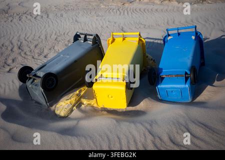 Drei bunte Kunststoff-Recyclingbehälter liegen auf den Seiten in den sandigen Stranddünen. Umgestürzte Müllcontainer, die Sturmschäden oder Abfallmanagement betonen Stockfoto