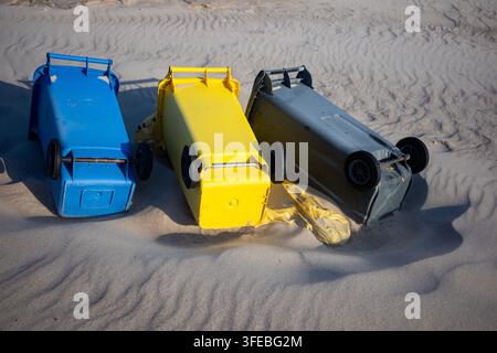 Drei bunte Kunststoff-Recyclingbehälter liegen auf den Seiten in den sandigen Stranddünen. Umgestürzte Müllcontainer, die Sturmschäden oder Abfallmanagement betonen Stockfoto