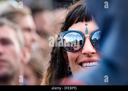 FESTIVALMENGE IN DER SONNENBRILLE EINER FRAU WIDERGESPIEGELT, REPORTAGE, GREEN MAN FESTIVAL 2019, BRECON, WALES, UK, 2019: Eine Nahaufnahme einer lächelnden jungen Frau mit Dreadlocks und Gesichtsjuwelen, ihre Sonnenbrille spiegelt die Festivalmenge und die Bühne vor ihr wider. Foto: Rob Watkins. INFO: Das Green Man Festival begann 2003 in Wales. Jährlich findet es am dritten Wochenende im August in den Brecon Beacons statt und feiert alternative Musik, Kunst, Literatur, Wissenschaft und familienfreundliche Kultur in einer wunderschönen Berglandschaft. Stockfoto