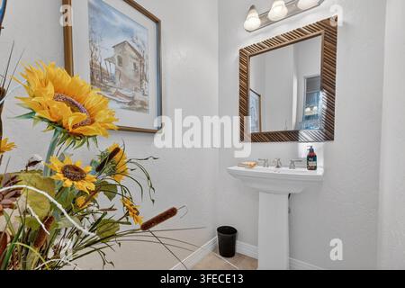 Helle Badezimmer-Szene mit lebhaftem Sonnenblumenstrauß im Vordergrund, großem gerahmten Gemälde an der Wand und holzgerahmter Spiegelfront über weißem Pedestalwaschbecken und Stockfoto