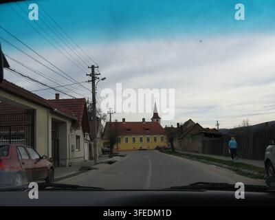 Blick aus einem Fahrzeug, das durch eine friedliche europäische Dorfstraße mit traditionellen Häusern und einem Kirchturm fährt. Stockfoto