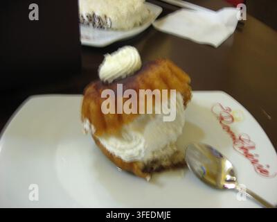 Ein traditionelles Rum Baba-Gebäck, gefüllt mit reichhaltiger geschlagener Sahne, serviert auf einem weißen Teller mit einem Teelöffel auf einem hölzernen Cafétisch. Stockfoto