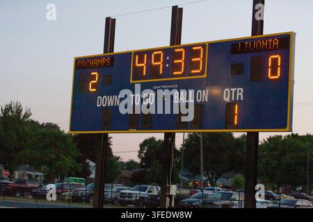 Eine elektronische Fußballstadion-Tafel im Freien, die in der Dämmerung beleuchtet ist, mit geparkten Fahrzeugen im Hintergrund. Stockfoto