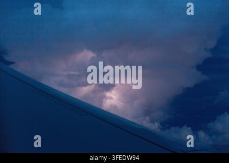 Blick aus dem Flugzeugfenster, der den Flügel vor einem dunklen Nachthimmel mit beleuchteten Gewitterwolken und einem dramatischen Blitzschimmer im Gewitter zeigt Stockfoto