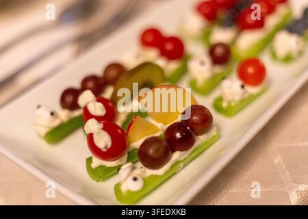 Kreative lustige Snacks aus Selleriestangen, gefüllt mit Frischkäse und belegt mit Trauben, Tomaten, Kiwi und Süßigkeitenaugen auf einem weißen Teller. Stockfoto