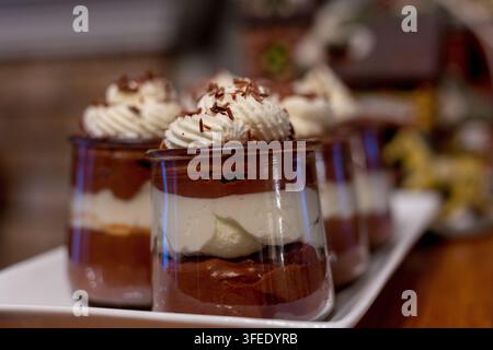 Schichtige Schokoladen-Mousse-Desserts, die in kleinen Gläsern serviert werden, mit Schlagsahne und Schokoladenraspeln auf einem weißen Tablett. Stockfoto
