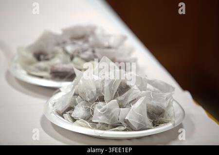 Haufen durchsichtiger Pyramiden-Teebeutel, gefüllt mit losen Teeblättern, serviert auf weißen Tellern auf einer Tischplatte. Stockfoto