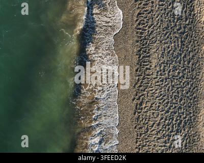 Drohnenaufnahme von oben, die tiefgrünes Meerwasser auf der linken Seite zeigt, das auf einen strukturierten Sand- und Kiesstrand mit weißer Schaumlinie trifft. Stockfoto